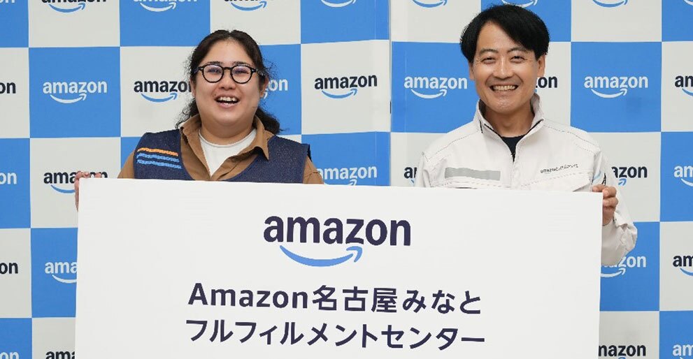 青と白の市松模様の壁の前で、白い作業着の人物と、茶色いシャツに紺色ベストの人物が、笑顔で大きなボードを持っている。ボードには「Amazon名古屋みなとフルフィルメントセンター」の文字。