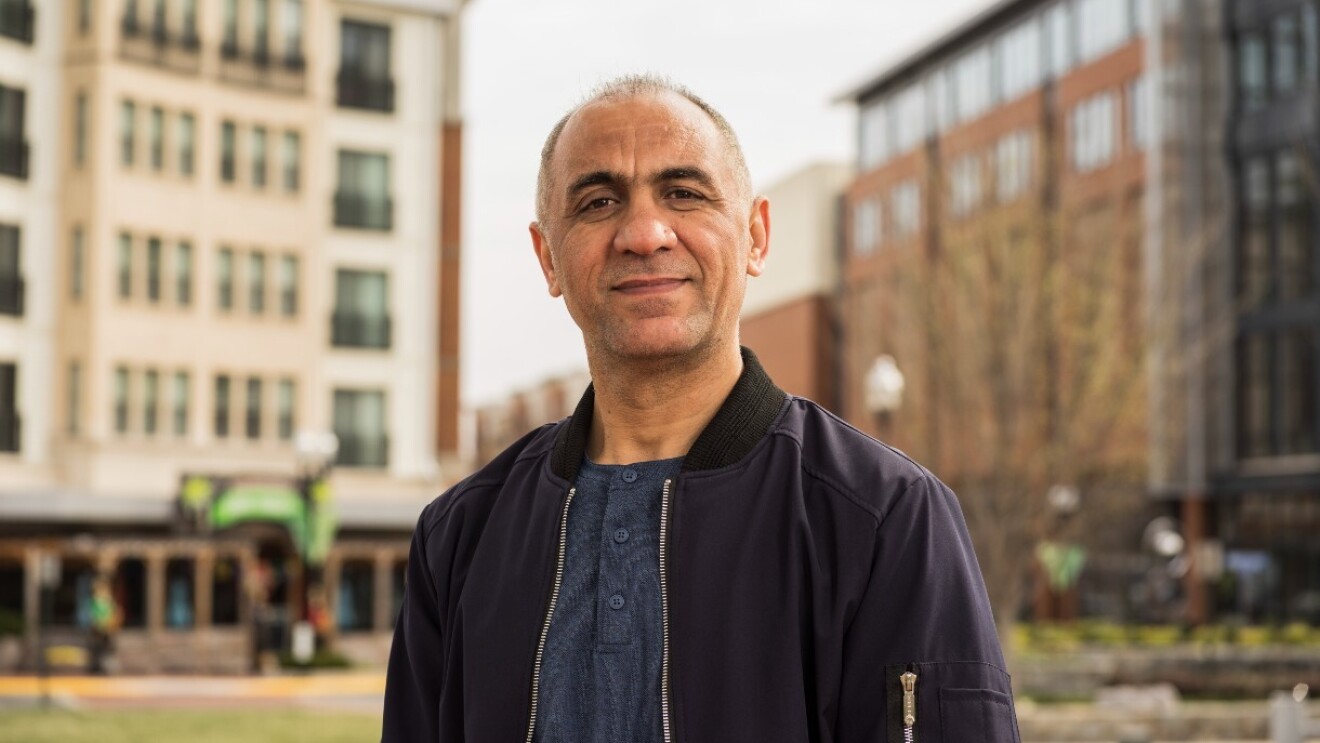 Khalid Noorzad stands outside in a park between buildings and grins at the camera.