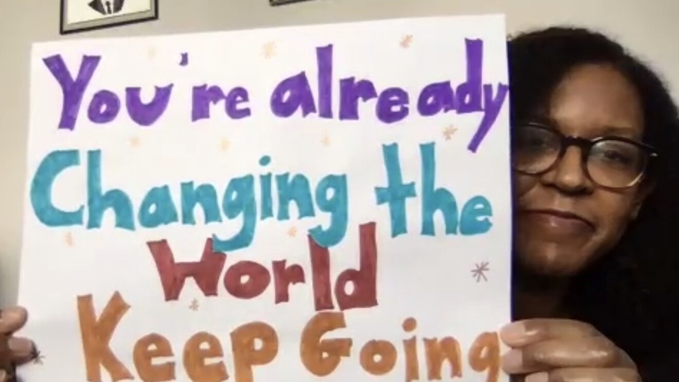 An image of a woman on a video call holding up a hand-drawn sign to her camera that says "You're already changing the world. Keep going." There are women on the call with her and their faces are in the smaller boxes at the top of the screen.