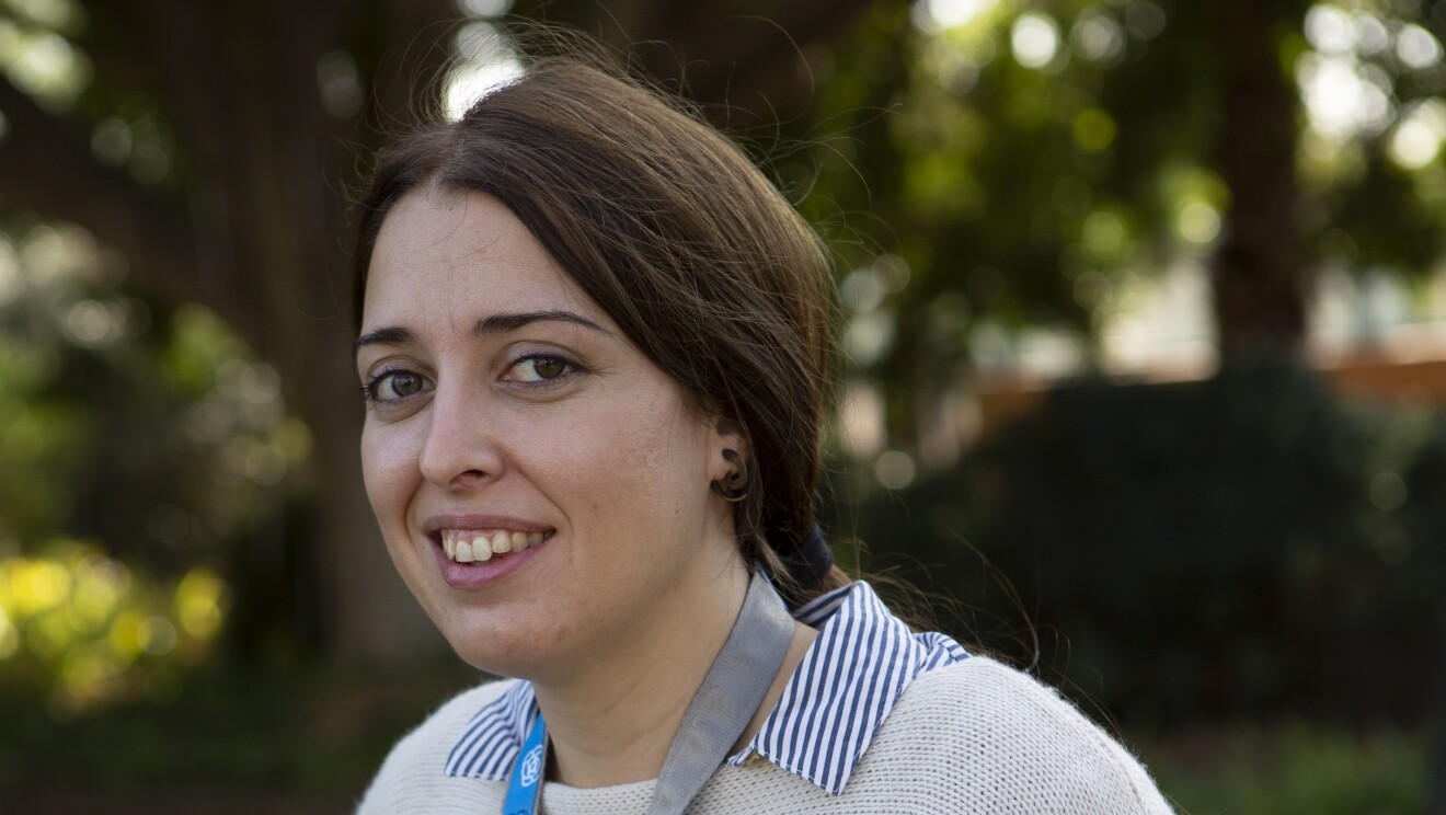 Alba Díaz es la protagonista de la foto. Mira a cámara y sonríe con los brazos detrás, en la espalda. Lleva el pelo largo y recogido en una coleta baja. Va con unos jeans, una camisa con rayas azules y blancas y un jersey de punto de color blanco roto con un cactus de color gris. La foto es exterior. 