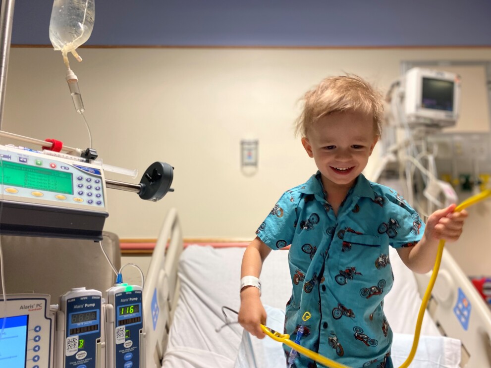 An image of a young boy smiling while standing up on a hospital bed playing with a yellow stethoscope.