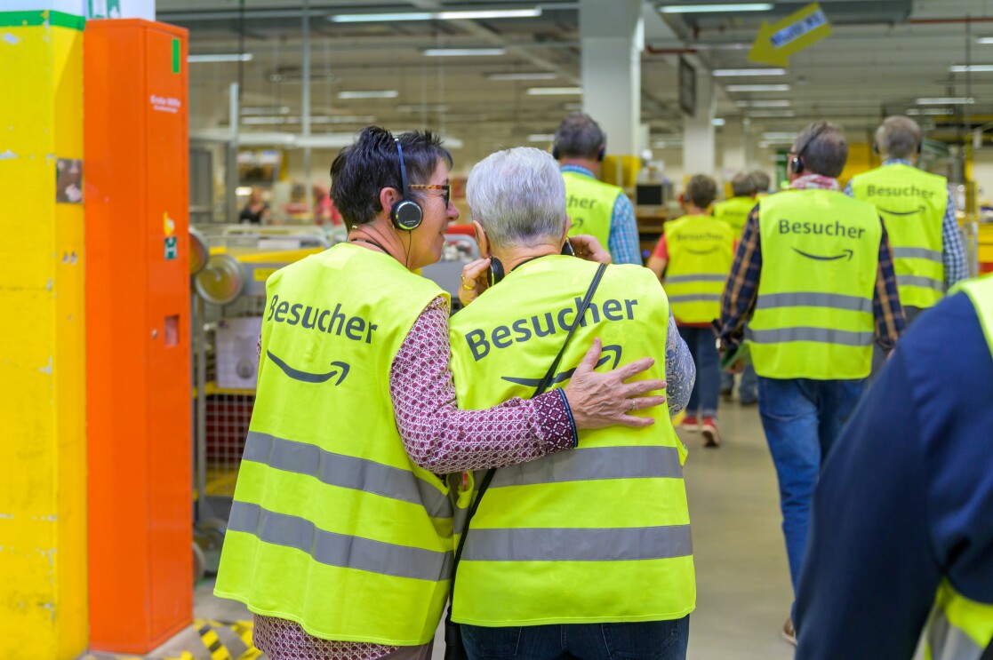 Zwei Besucherinnen in gelben Sicherheitswesten von hinten: Auf den Westen steht "Besucher" und das Amazon Smile Logo ist zu erkennen.