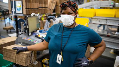 An Amazon associate working at a fulfillment center pack station poses for the camera while wearing a protective surgical mask.
