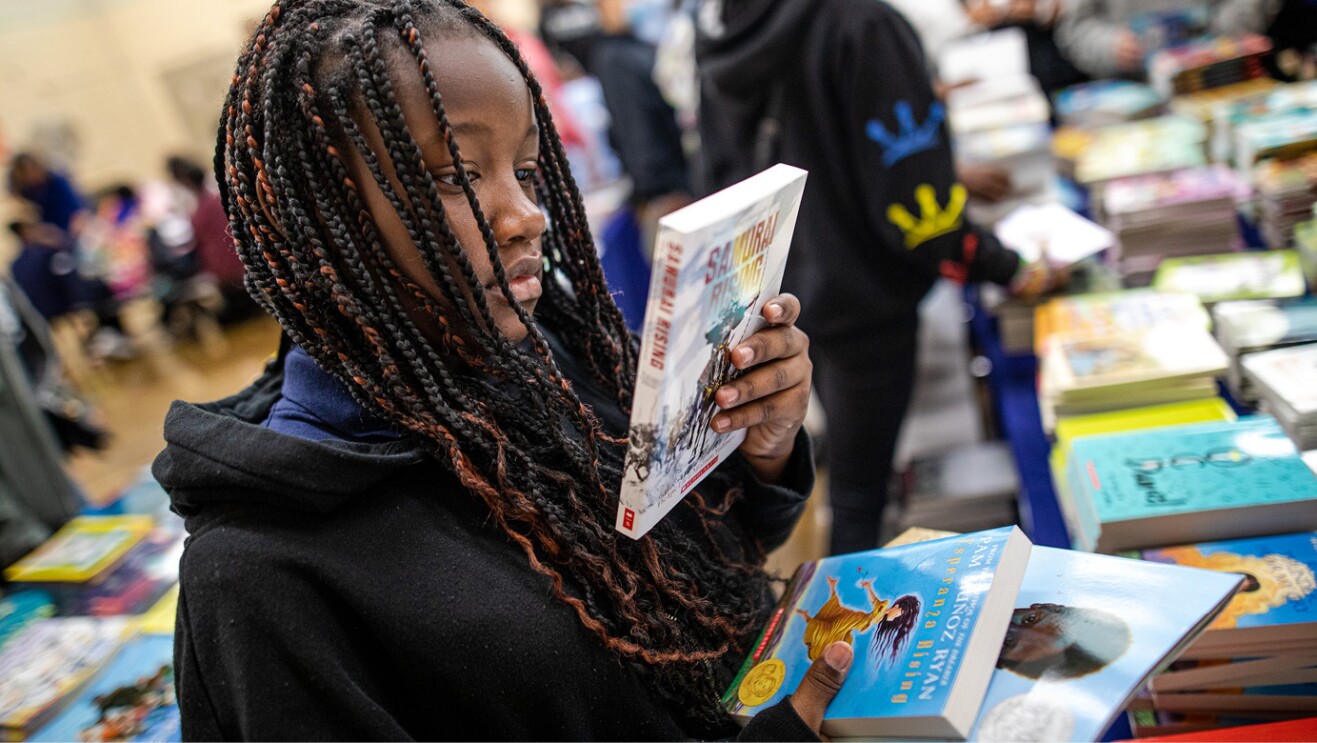 An image of a girl looking at a book while holding other books in her other hand.