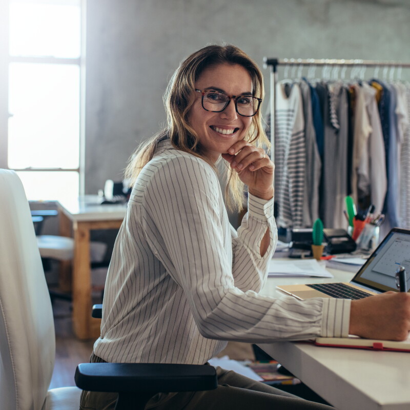 En un taller con una burra de ropa en el fondo con ropa colgada, hay una mujer sentada en una mesa. De fondo se ve un ventanal grande de color blanco y tres meses y dos sillas. En el centro de la foto hay una mujer sentada en una silla con una agenda y un ordenador delante. La mujer mira a cámara sonriendo. Lleva gafas de pasta, una camisa blanca con rayas negras diplomáticas. Con la mano izquierda se aguanta la cara y la mano derecha agarrango un bolígrafo escribiendo en la agenda. 