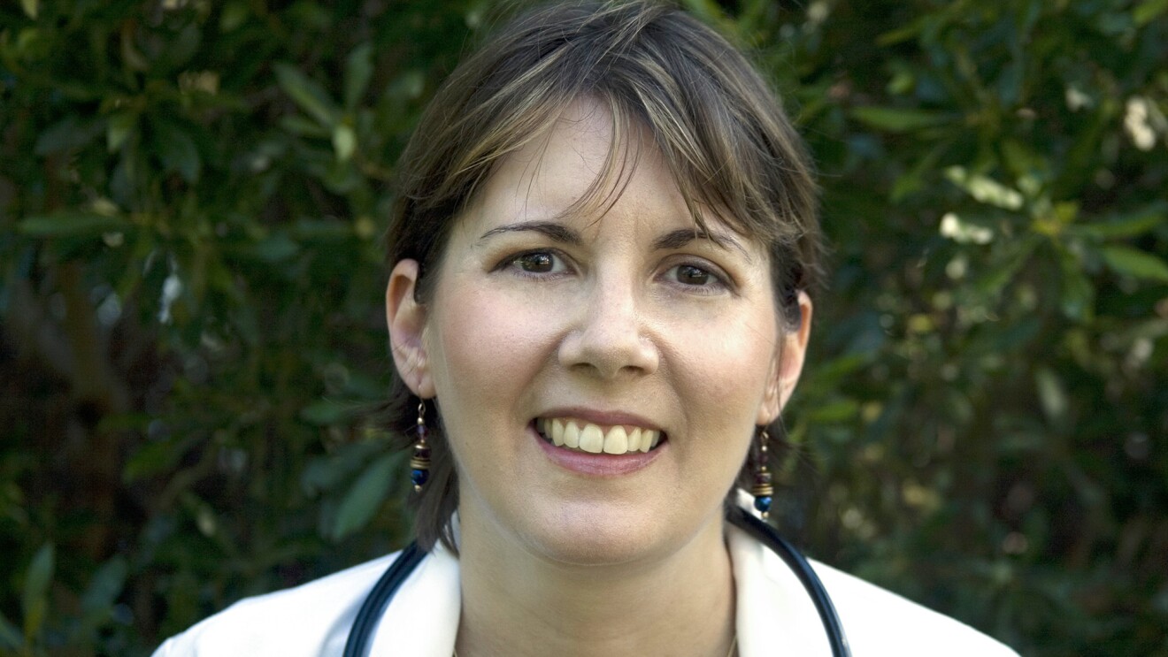 亚马逊作者CJ里昂笑到相机。她头发乌黑,穿着一件白色夹克,脖子上听诊器。背景是模糊树。