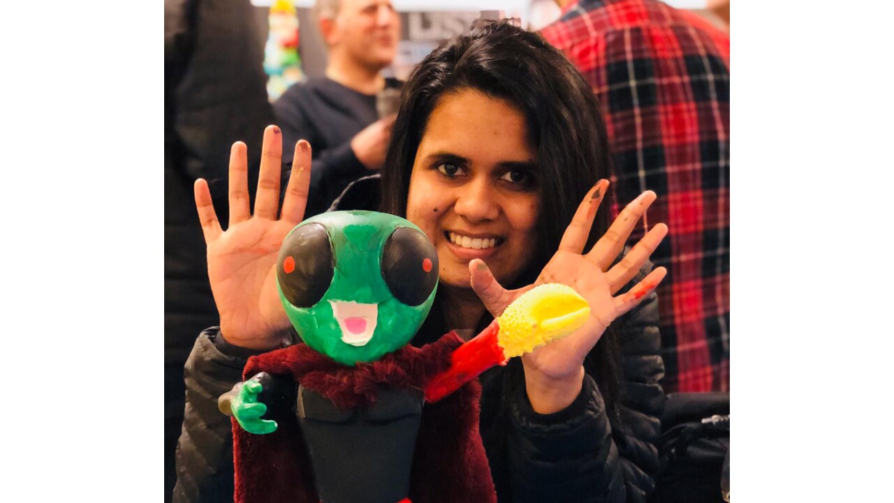 An image of a woman smiling for a photo holding both of her hands in front of her. There is an alien figurine in front of her and paint on her fingers indicating she painted the figure.