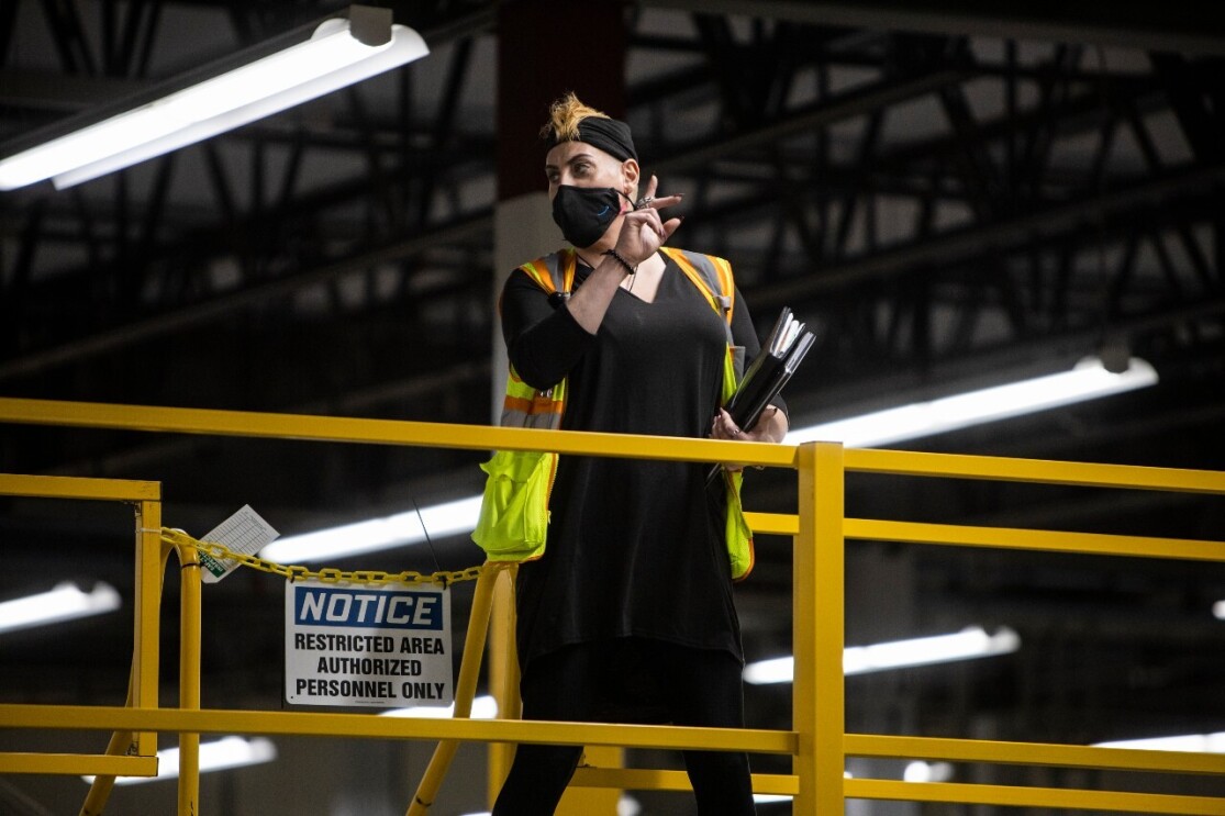 An image of an amazon employee standing in a fulfillment center wearing a mask and a safety vest while giving instructions to fellow employees.