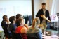 Amazon Campus Challenge. En una aula de ofrmación hay un candidato de abirendo un sobre y después una mesa con cinco personas de espaldas a nosotros mirando al candidato. El candidato lleva americana azul y pantalones chinos claritos. Sujeta con una mano un sobre de cartón y con la otra está intententando sacar lo que hay dentro. 