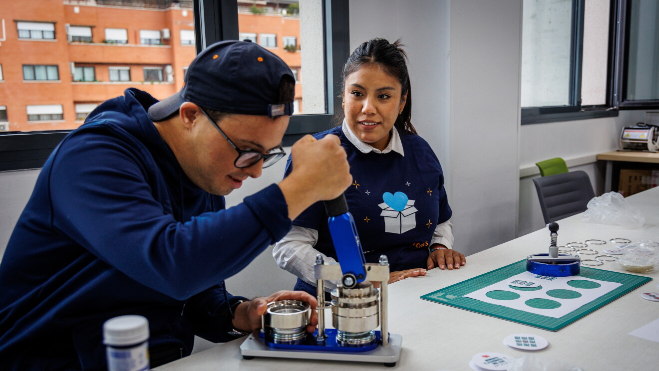 En las instalaciones de Down Madrid en Madrid aparece una participantes de la asociación y un trabajador de Amazon. La trabajadora van co la misma camiseta azul y el badge colgado y el de Down Madrid está cortando con una máquina unos papeles redondos que son una prueba de la gincana. 