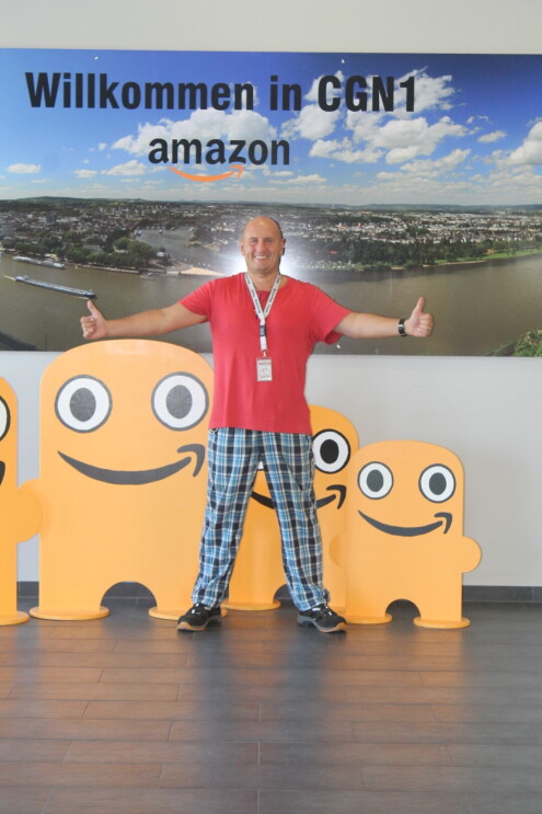 Ein Amazon Mitarbeiter in karierter Schlafhose und rotem Shirt steht mit ausgebreiteten Armen vor einer Fotorückwand mit Aufschrift "Willkommen in CGN1"