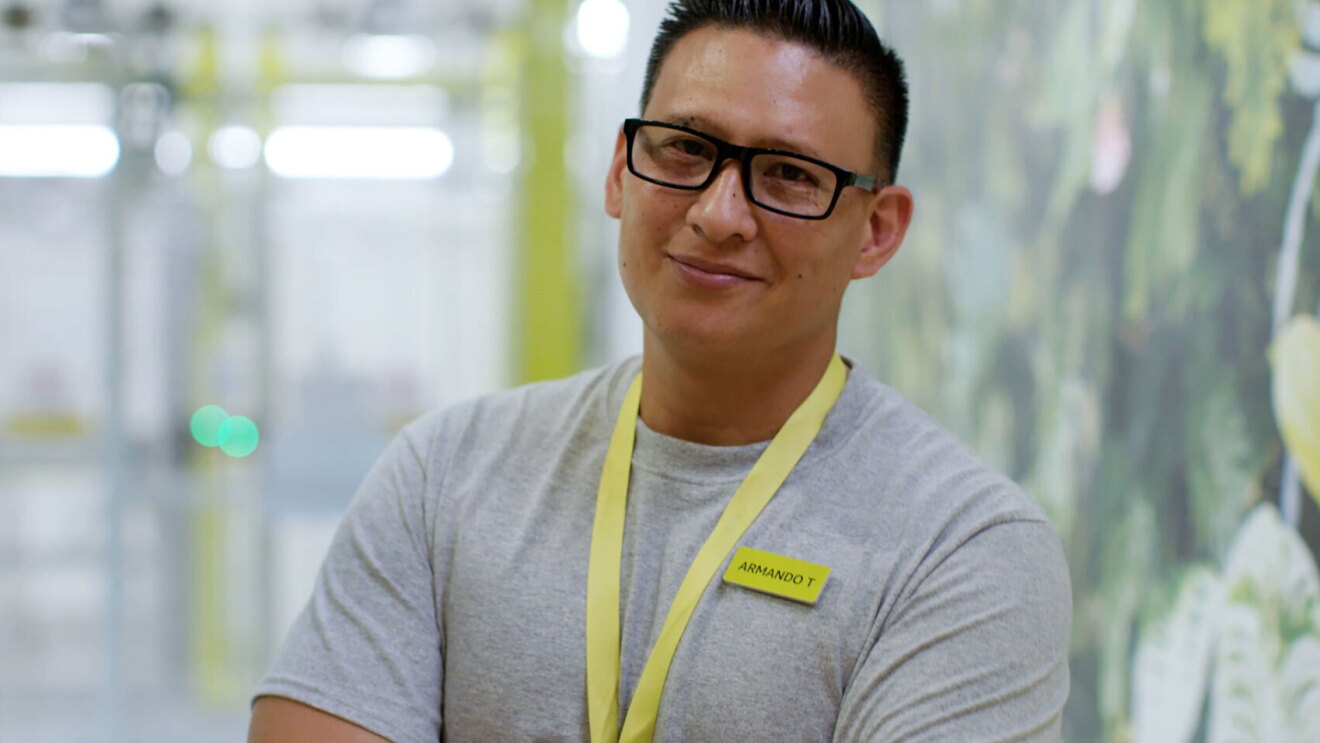 An image of a man smiling for a photo while standing in an Amazon Style store. He is wearing a neon yellow lanyard and a neon yellow name tag. 