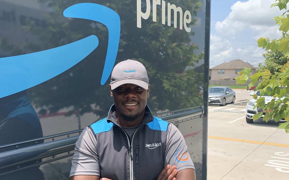 An image of a man standing in front of an Amazon delivery truck with the Amazon logo and the words "Prime" written on the side. He is wearing an Amazon-branded hat and vest and smiling with his arms folded in front of him.