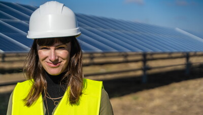 Denise Hugo, jefa de proyectos de Energías Renovables de Amazon. Ella está delante de las placas solares. Va vestida con casco blanco y chaleco amarillo. Tiene el pelo castaño y le llega más allá de los hombros. LLeva tres collares de oro que están por encima de un jersey de cuello alto marrón.  