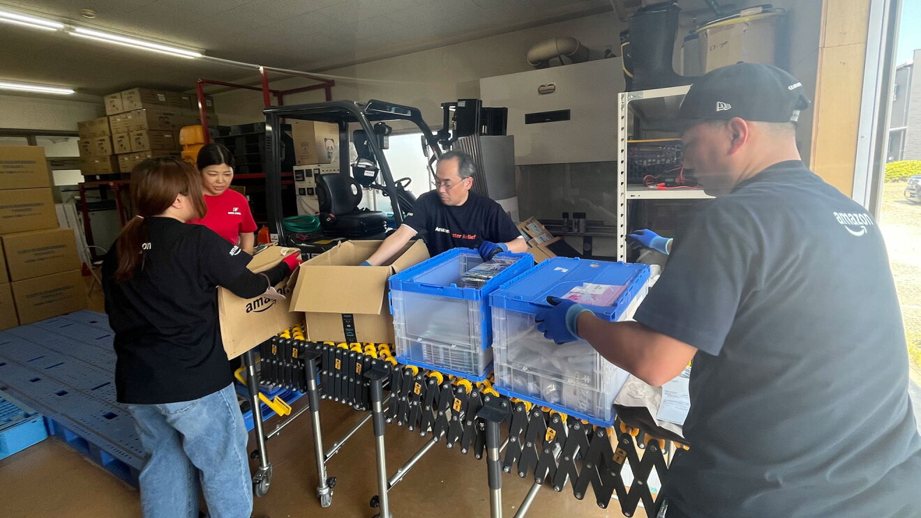 段ボールや資材が多く積まれた作業場で、荷物の箱を開けて作業をしている4人の人物。手袋を着用している。