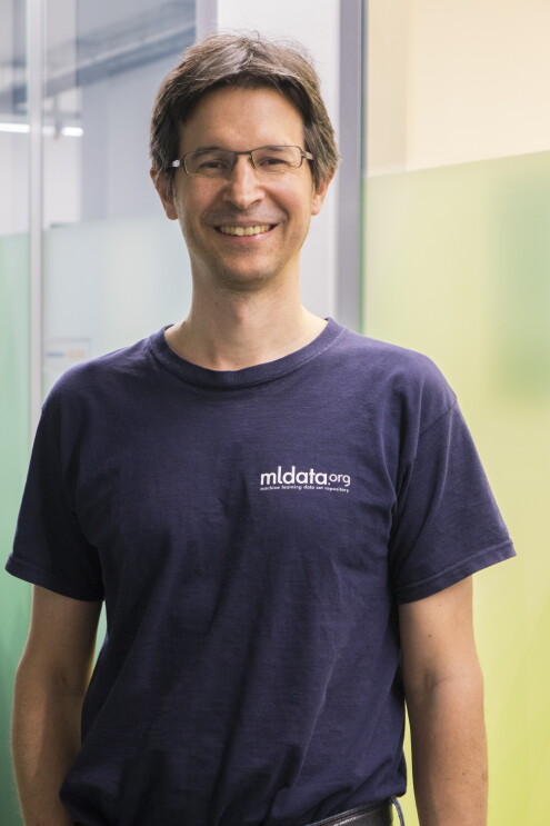 Ein Mann mit braunen Haaren, Brille und einem blauen T-shirt lacht in die Kamera.