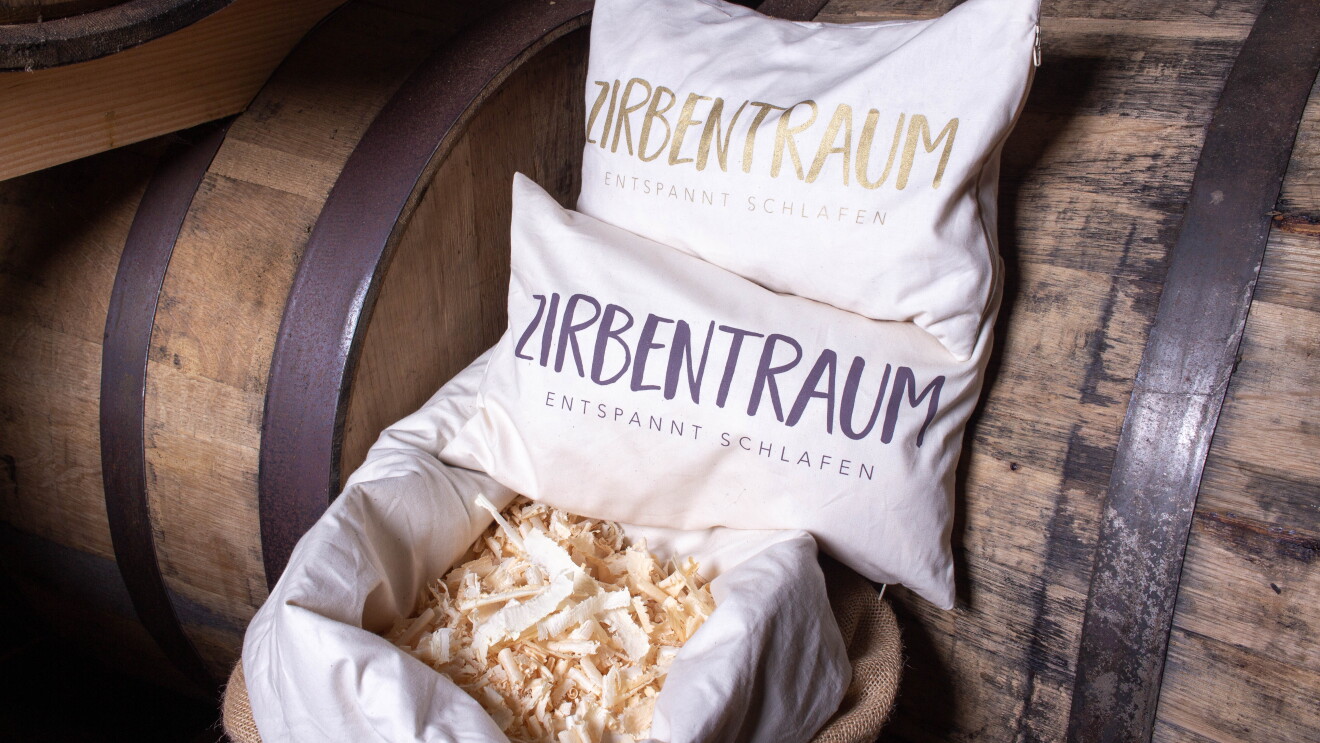 Zwei weiße Kissen mit der Aufschrift Zirbentraum liegen auf Holzfässern.