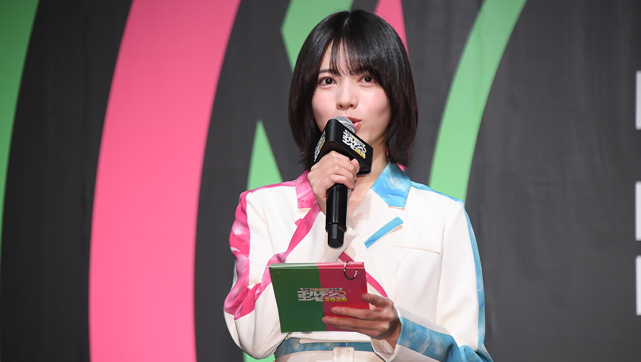 黒い背景にピンクと緑の曲線が入った壁の前に立つ、若い女性の上半身。白地にピンクと水色のアクセントが入ったスーツを着て、右手にマイク、左手に冊子を持っている。