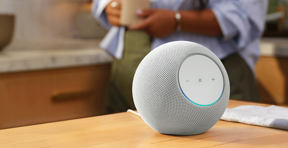 キッチンのテーブルに白い球状のデバイスが置かれている。デバイス本体は全体的にメッシュ状、前面に丸く白いパネルがある。テーブルの後ろに人物が立っている。