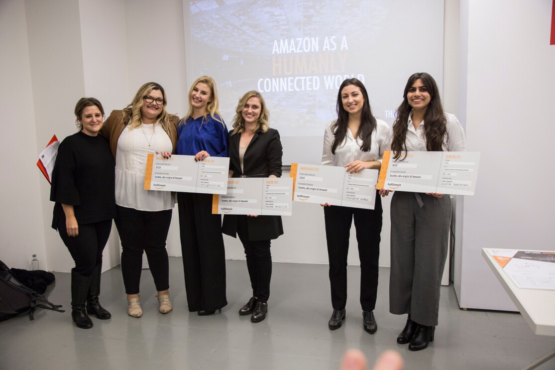 Sei ragazze in posa mentre reggono dei biglietti aerei giganti. Quattro di loro sono Monika, Urte, Francesca e Ankita, quattro studentesse del Master in Interior Design dello IED Milano che hanno vinto gli Amazon Design Awards, una competizione in cui si chiedeva ai corsisti di ripensare i depositi di smistamento Amazon con uno sguardo alla praticità e al comfort dei dipendenti. Sullo sfondo, uno schermo di proiettore. 
