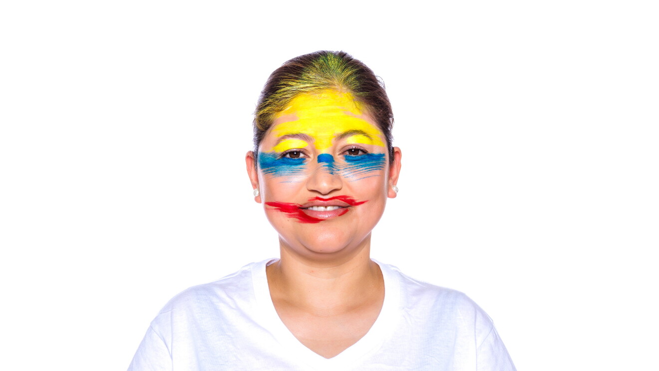 Para el Día de la diversida Amazon ha preparado una acción con asociados de diferentes países que llevan la bandera pitnada en su cara. En la foto, Gladys con camiseta blanca, mirando sonriendo a cámara con la bandera de su país puntada en la cara. Tiene el pelo marrón y una coleta detrás. Lleva pendientes en las orejas. 