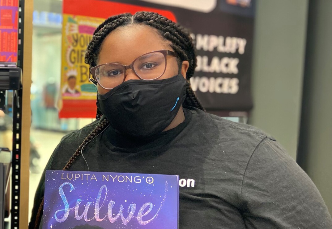 An image of a woman standing in an Amazon bookstore smiling for a photo under her mask. She is holding a children's book titled "Sulwe," authored by Lupita Nyong'o. 