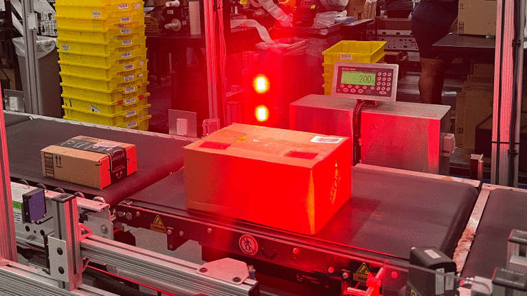 An image of a box on a conveyer belt. There is a big red light flashing on it.
