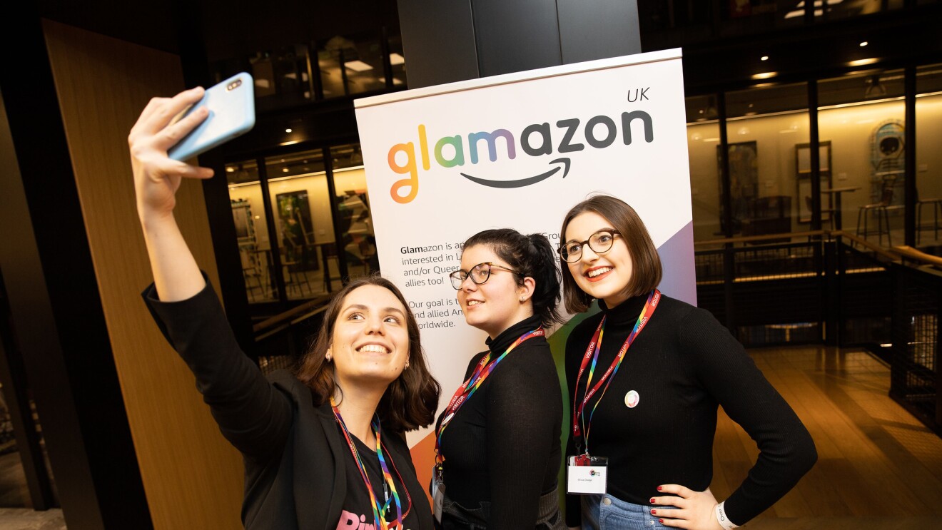 Two Amazon employees taking a selfie with a representative from PinkNews at the 2020 British LGBT+ Awards. They are standing in front of a glamazon banner.