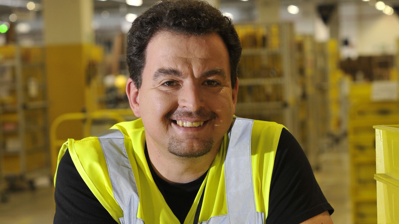 Nikolai Lisac, ein Mann mit Sicherheitsweste und braunem, gelocktem Haar, lehnt locker an einen Stapel von gelben Behältern in einem Logistikzentrum