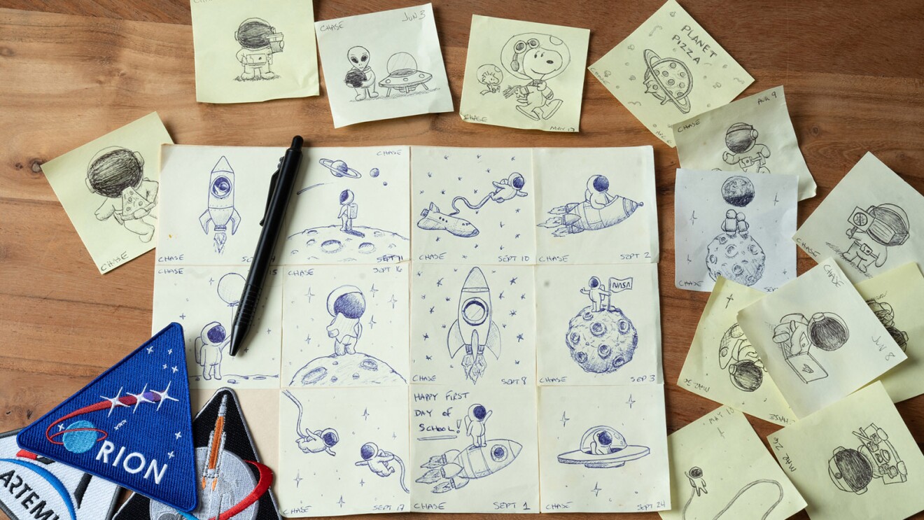 An image of a desk with a notepade on it and sticky notes surrouding it. On the notepad and sticky notes are drawings of rocket ships and astronauts in space.
