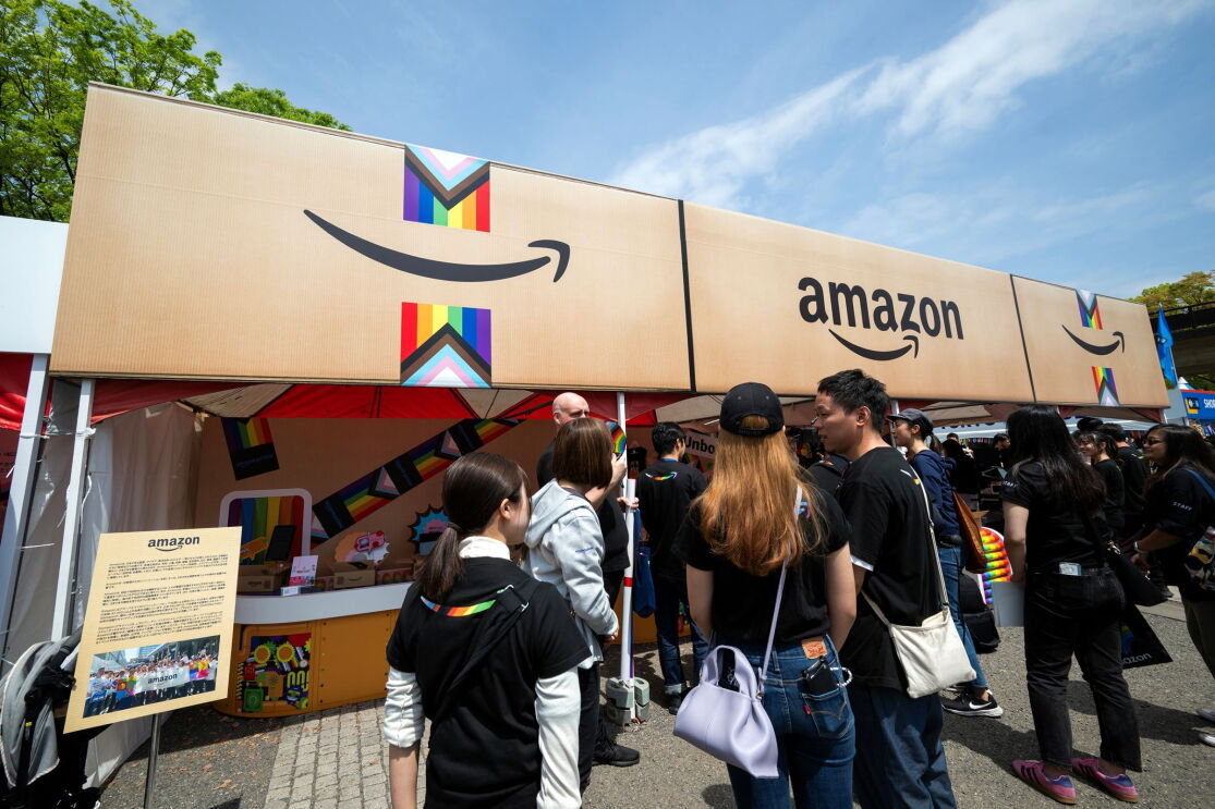 屋外のイベントブース。レインボーカラーのテープが貼られたAmazonの段ボールを模した屋根の下に人々が並んでいる