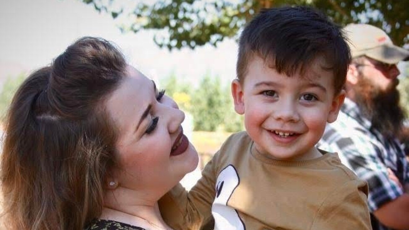 An image of a mother holding her son while looking up at him and smiling. Her son is also smiling and they are outside at a family gathering.