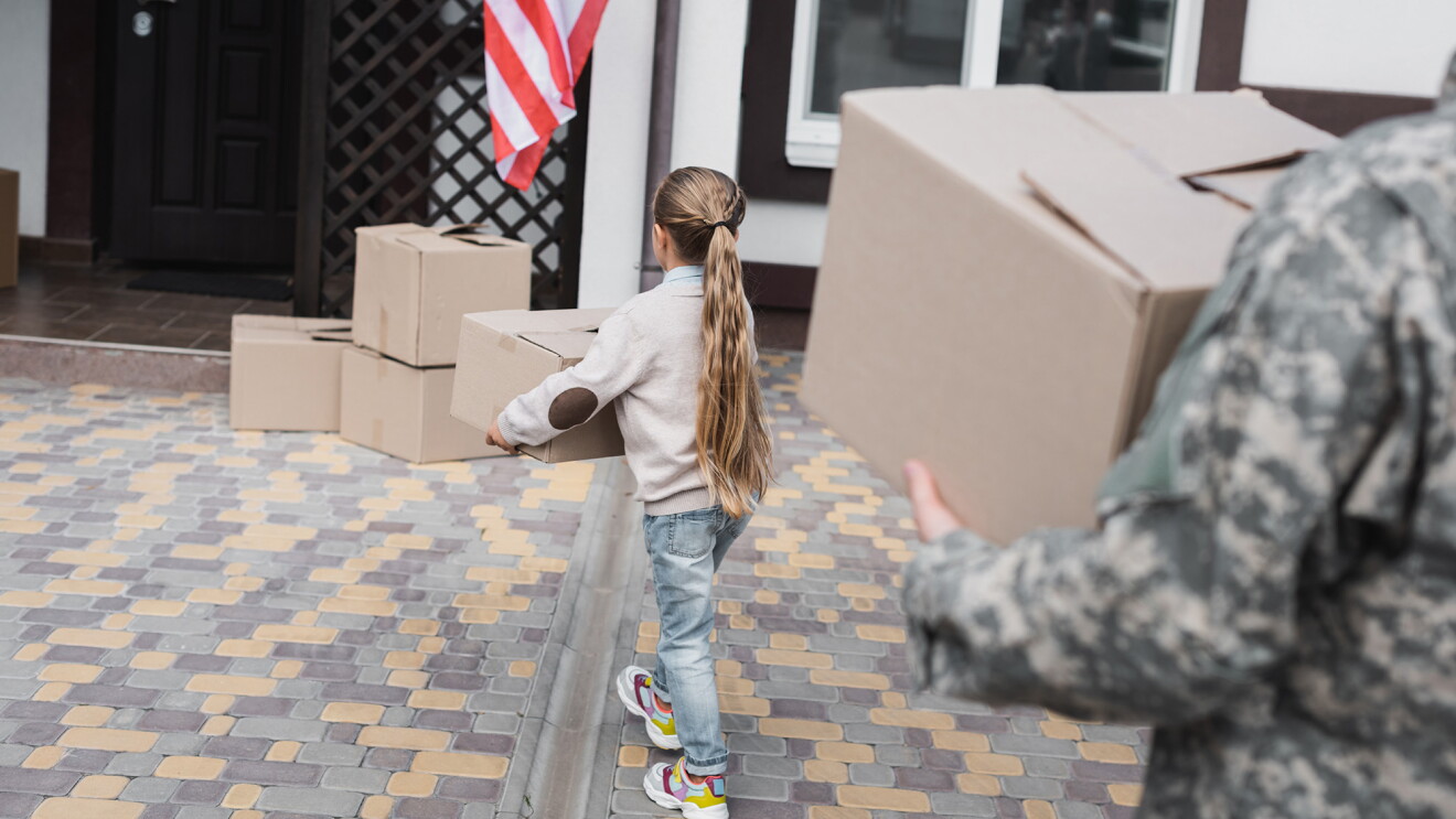 A child carries a moving box, behind her a man in military fatigues also carries a box. 