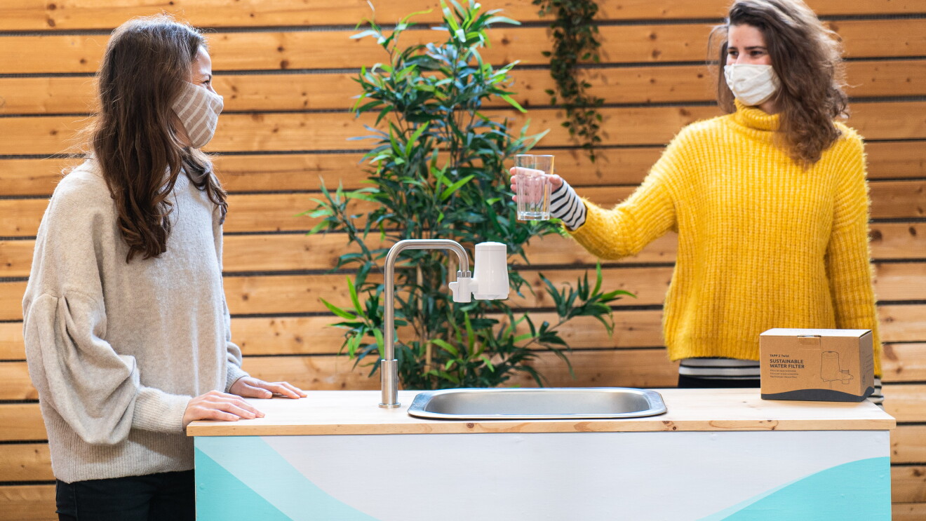 La start-up TAPP Water fue fundada por Magnus Jern y Alexander Schwarz. En la foto aparece la CEO Rocío Alcocer con una compañera de trabajo. Las dos están en una mesa que tiene un grifo con el filtro. Rocío va con una mascarilla de color marrón con rayas blancas y un jersey también marrón muy claro. Unos pantalones negros y tiene el pelo largo castaño y las manos apoyadas sobre la mesa. Su compañera va con un jersey de color amarillo el pelo castaño y ondulado. Sujer una vaso de cristal con agua. Encima de la mesa una caja con el filtro de agua. De fondo una pared de madera.  
