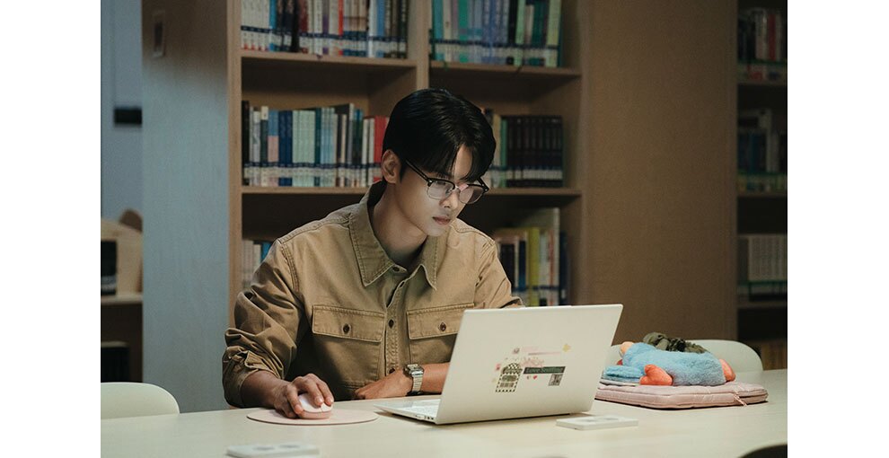 図書室で机に向かってノートパソコンを使う眼鏡の男性。右手でマウスを操作し、画面を見つめている。