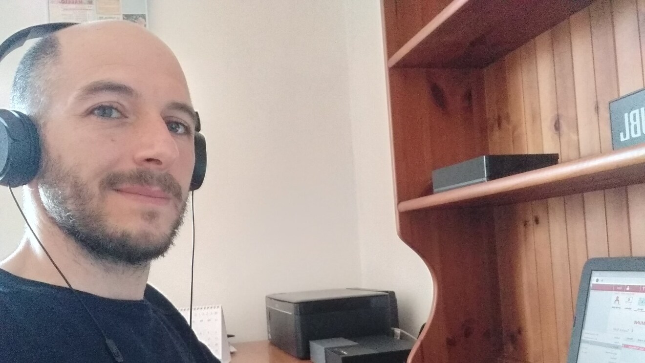 Primo piano di un uomo sulla sinistra. Indossa una maglia nera e delle cuffie, siede di fronte a una scrivania su cui è posato un PC.