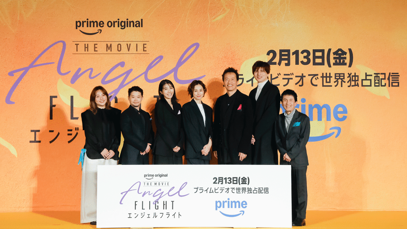 オレンジ色の壁の前、オレンジのステージの上に、黒い服を着て横一列に並んだ7人の人物。右端の人物だけグレーの上下。人物たちの足元に長方形のパネルが置かれている。パネルには「エンジェルフライト」の文字。