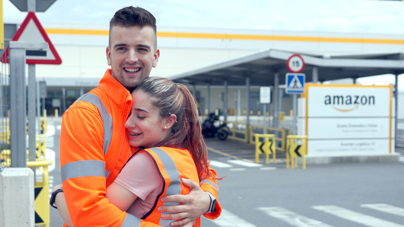 En la foto aparecen Amanda y Pedro, dos trabajadores del centro logístico de Amazon de Illescas (Toledo) después de la pedida de mano. Están abrazados, él le pasa la cabeza, Están situadas en el aparcamiento exterior del centro logístico. En el margen derecho aparece el cartel de Amazon. Él va con un chaqueta naranja con las badas grises y abraza a Amanda, que lleva una camiseta de manga corta rosa y un chaleco también naranja. Ella lleva dos piercings en las orejas. Ella va con el pelo recogida en una cola que le llega más allá del hombro y el lleva el pelo corto y barba de dos días. 