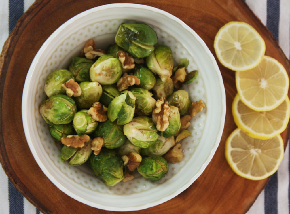 An image of brussels sprouts with walnuts on them in a plate sitting on a cutting board with lemon slices next to them.