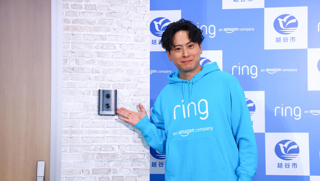 ダミーの壁に取り付けられたインターホンを手で指し示す男性。ringロゴ入りのパーカーを着用