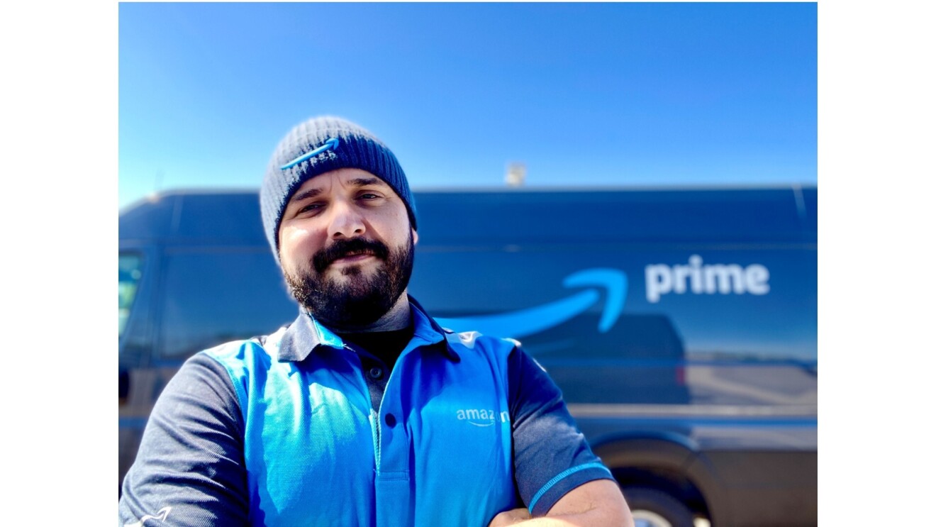 Nick Gallina smiles for the camera in front of his Prime delivery van. He is wearing a blue collared T-Shirt with an Amazon logo and a blue beanie with an Amazon logo.