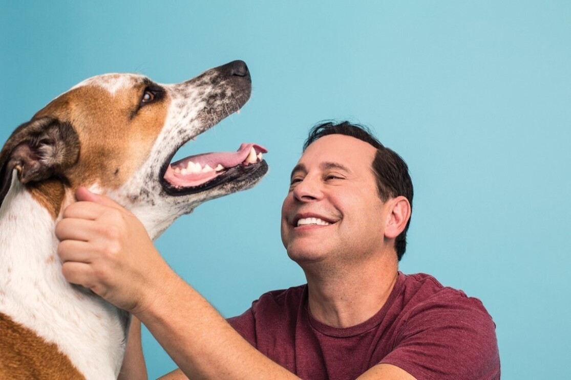 An image of a man smiling while looking at and petting his dog. It is a professional photo against a blue background.