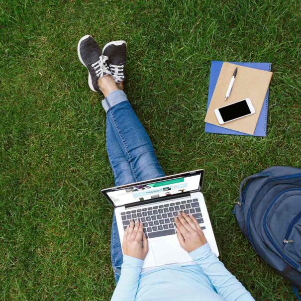 A woman types on the keyboard of a laptop while sitting in the grass.