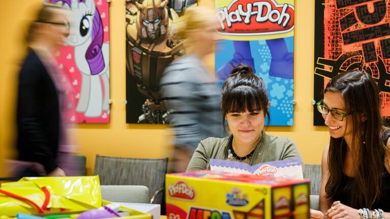 Two women in the foreground sit at a table surrounded by packaging. Other people pass by in the background. The wall has posters of My Little Pony, Play-Doh, and other Hasbro brands.