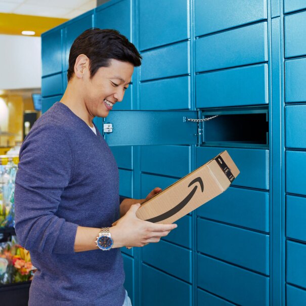 An Asian man in a blue sweater picks up his Amazon package at a Locker location. Colorful flower bouquets are in the background.