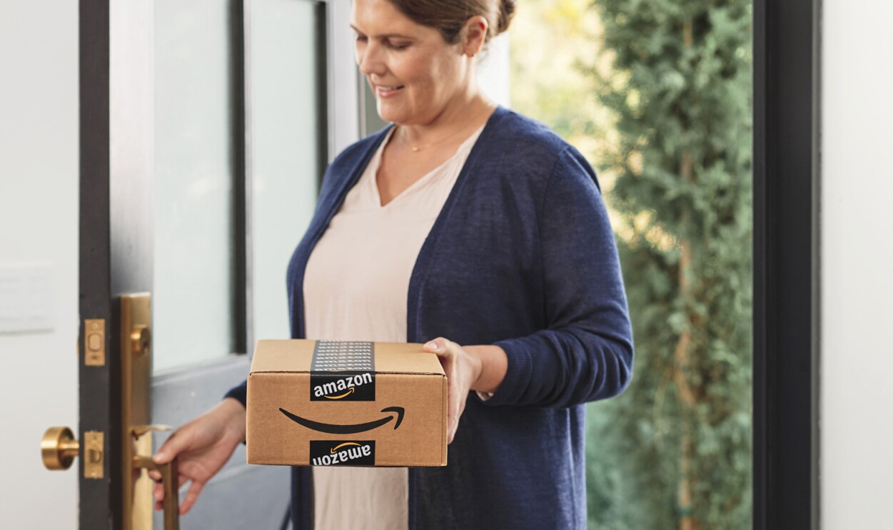 A woman walks in the front door of a home carrying an Amazon package.