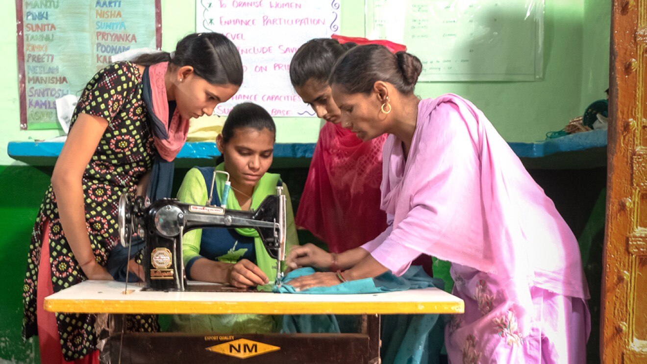 Three women stand around a woman who sits at a sewing machine.