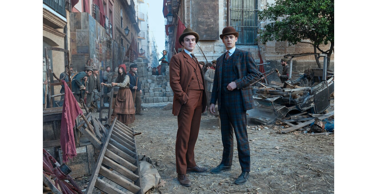 がらくたや廃棄物が放置された空き地に立つ、身なりのいいスーツ姿の男性2人。2人の背後には労働者階級らしき人々。