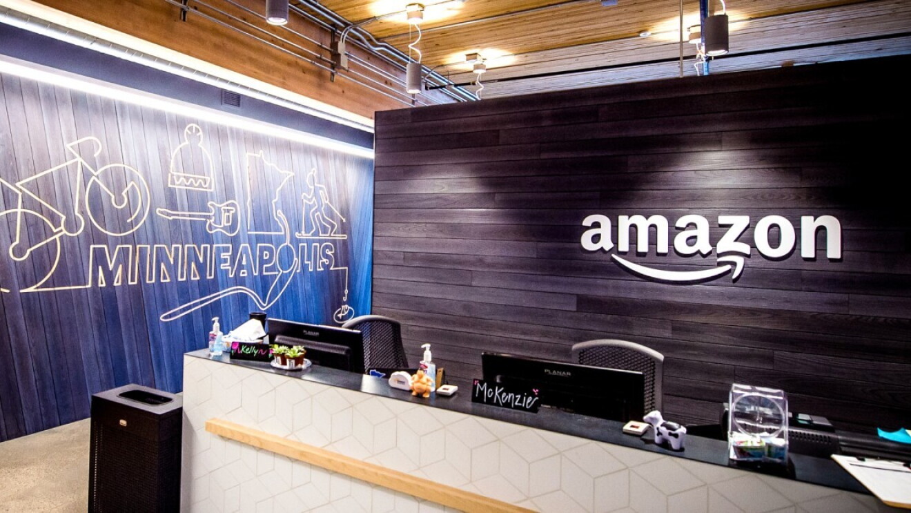 Z przodu recepcja, w tle ścianka stylizowana na drewnianą z białym logo Amazon, z boku w tle ścianka stylizowana na drewnianą z tekstem "Minneapolis" oraz ikonkami roweru, czapki, gitary, triatlonisty oraz konturem granicy stanu