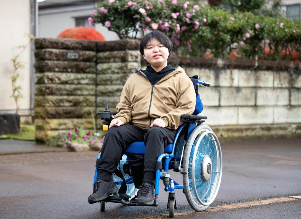 青い車椅子に座っている男性が道の脇に止まっている。彼の後ろはレンガのフェンスの後ろからピンク色の花が咲いている。